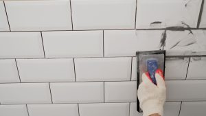 Grouting seams on a tile backsplash. Workers' hands using a rubber spatula and grouting with paste between white ceramic tiles. Close-up of grout.